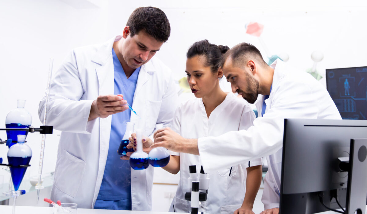 Team of scientist in white coat working together with smoking blue liquid in modern research lab.